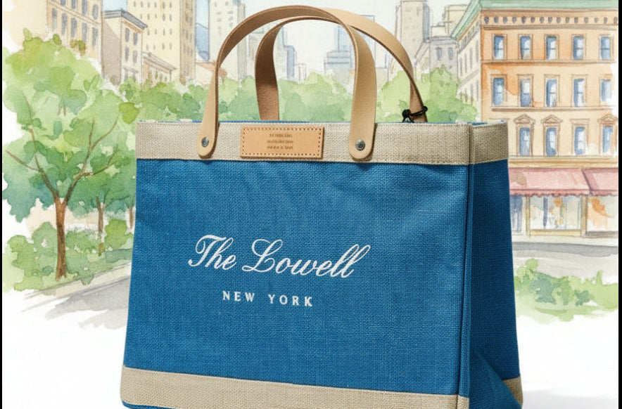 A canvas tote bag from The Lowell New York featuring a blue front panel, beige trim, and tan leather handles, set against a watercolor-style city park backdrop.